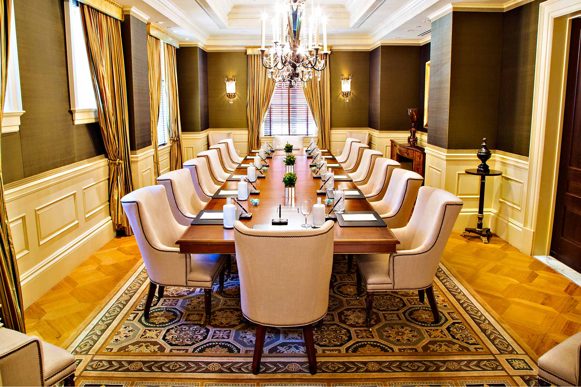 The Parlor Boardroom is an impressive meeting space designed for up to 26 Participants around a grand wood polished table crowned by tall taper-like chandeliers.